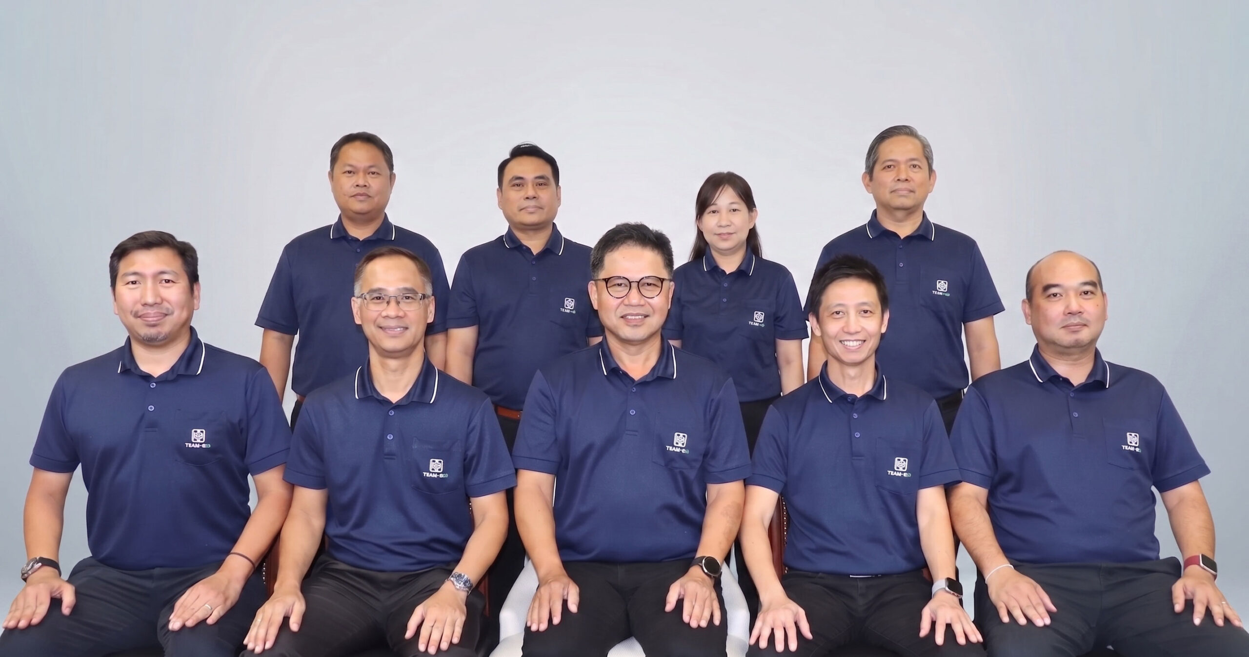 คณะผู้บริหาร บริษัทบริหารโครงการก่อสร้าง TEAM CM ทั้ง 9 ท่าน สวมเสื้อโปโลสีน้ำเงินสกรีนโลโก้บริษัท ยืนและนั่งถ่ายภาพร่วมกันบนพื้นหลังสีเทา